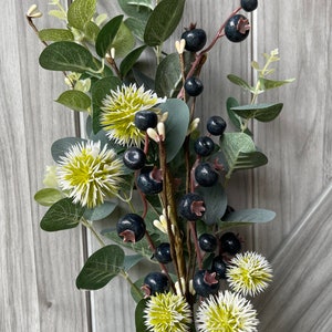 May include: A decorative floral arrangement featuring dark blue berries, green eucalyptus leaves, and spiky white and yellow flowers. The arrangement is set against a gray wooden background, creating a natural and rustic aesthetic.