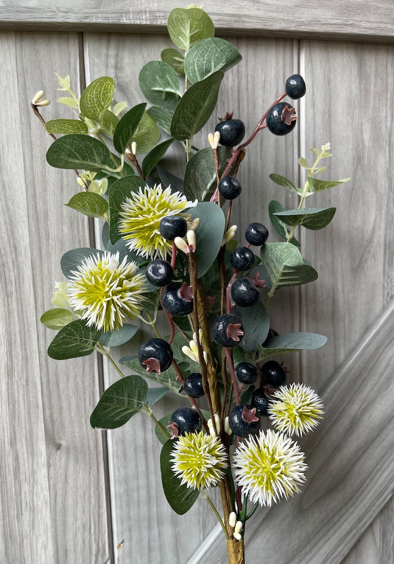 May include: A decorative floral arrangement featuring dark blue berries, green eucalyptus leaves, and spiky white and yellow flowers. The arrangement is set against a gray wooden background, creating a natural and rustic aesthetic.