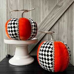 May include: Two decorative pumpkins featuring alternating segments of bright orange velvet and black and white checkered fabric. Each pumpkin is topped with a glittery silver stem and accented with vertical rows of clear rhinestones between sections.