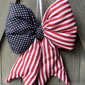 May include: A large decorative bow featuring a patriotic design. One side is navy blue with white stars, and the other side has red and white stripes. A white ribbon is attached for hanging. The bow is made of fabric and is suitable for festive decor.