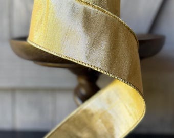 May include: A spool of metallic gold ribbon with a subtle textured finish and a shimmering sheen. The ribbon features a structured edge and is wound around a cylindrical core, displaying a continuous, flowing length of fabric with a reflective surface.