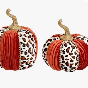 May include: Two decorative pumpkins featuring alternating vertical panels of orange velvet and white fabric with a brown and black leopard print. Each pumpkin is topped with a textured gold stem and accented with thin lines of clear rhinestones.