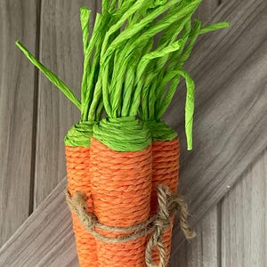 May include: Three orange woven carrots with green tops tied together with twine. The carrots are a decorative item, possibly for Easter or spring. The background is a wooden surface.