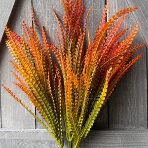 May include: A decorative artificial plant stem featuring multiple branches with small, oval-shaped leaves. The foliage displays a gradient of colors, transitioning from deep green at the base to vibrant orange and red at the tips of each branch.