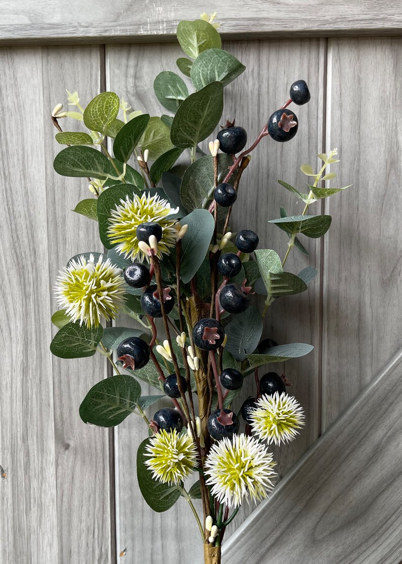 May include: A decorative floral arrangement featuring dark blue berries, green eucalyptus leaves, and spiky, light green and white flowers. The stems are brown and wrapped with a gold-colored material. The background is a light gray wooden surface.