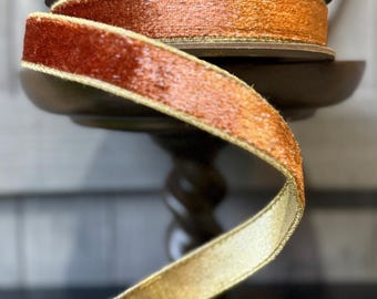 Könnte beinhalten: Eine Spule aus kupferfarbenem Samtband mit metallisch goldenem Rand. Das Band ist auf eine runde Spule gewickelt und verläuft spiralförmig nach unten, wobei die strukturierte Samtoberfläche und der schimmernde Goldrand beidseitig sichtbar sind.