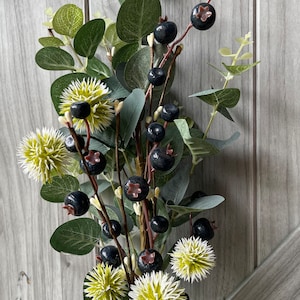 May include: Artificial floral arrangement featuring dark blue berries, green leaves, and spiky, light green and white flowers. The arrangement is set against a light gray wooden background, creating a natural aesthetic.