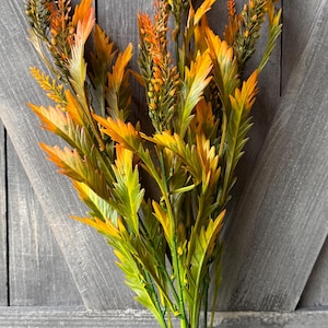 May include: A bundle of artificial flowers with green stems and a mix of yellow, orange, and green leaves. The flowers have a feathery appearance and are arranged against a gray wooden background.