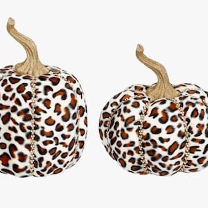 May include: Two decorative pumpkins featuring a white, brown, and black leopard print pattern. Each pumpkin has a textured gold glitter stem and vertical rows of small, clear, reflective beads separating the segments of the pumpkin surface.