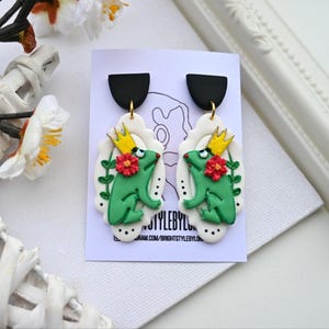 May include: A pair of green frog earrings with gold crowns and red flowers. The earrings are made of polymer clay and have a white background with black polka dots.