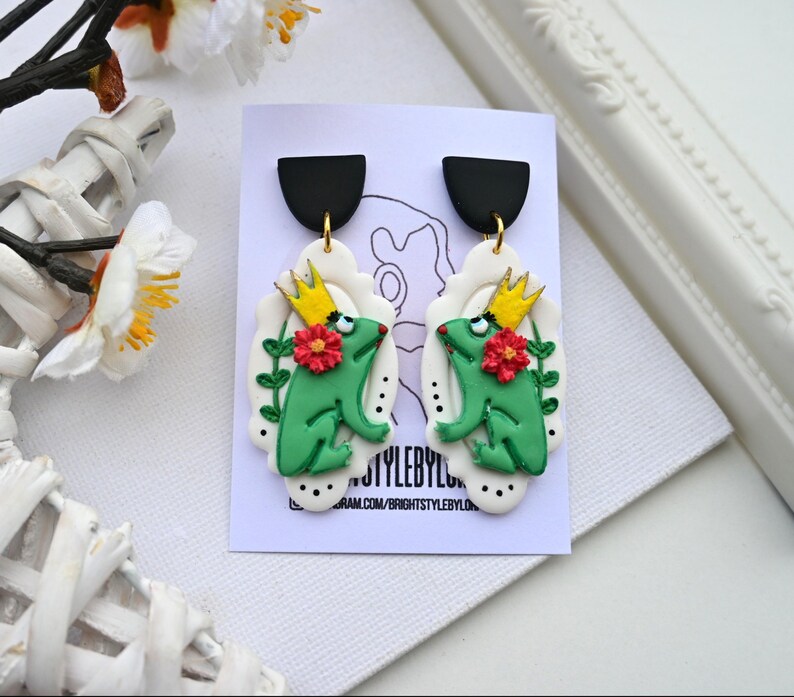 May include: A pair of green frog earrings with gold crowns and red flowers. The earrings are made of polymer clay and have a white background with black polka dots.