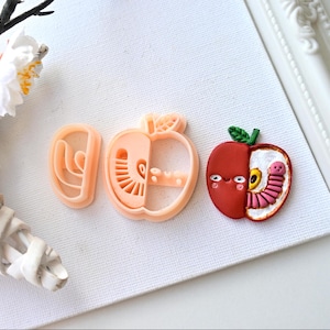 Peut inclure: Un emporte-pièce en forme de pomme et une pomme décorative. L'emporte-pièce est de couleur pêche et montre une coupe transversale d'une pomme. La pomme décorative est rouge avec une feuille verte et un ver souriant à l'intérieur.