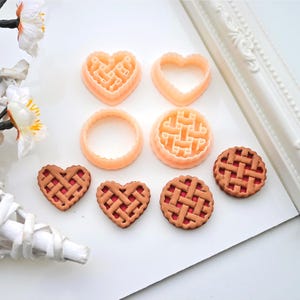 Pode incluir: Uma coleção de ferramentas de panificação e mini tortas. As ferramentas incluem cortadores de massa de torta em forma de coração, círculo e redondos. As mini tortas têm um design de crosta de treliça com um recheio vermelho. As ferramentas são de cor pêssego e as tortas são marrons e vermelhas.