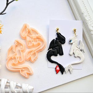 Op de afbeelding: Draakvormige oorbellen, één zwart en één wit, met goudkleurige oorbelpalen. Er is ook een bijpassende set draakvormige koekjessnijders. De oorbellen en snijders liggen op een wit oppervlak.