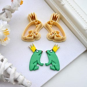 May include: Two green frog cookie cutters with gold crowns and a pair of matching frog cookie cutters in a light brown color.