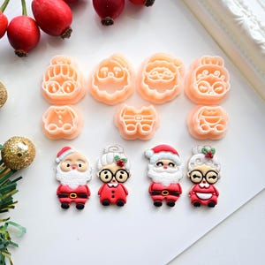 K&ouml;nnte beinhalten: Eine Sammlung von weihnachtlichen Ausstechern und dekorativen Figuren. Die Ausstecher haben die Form von Gesichtern des Weihnachtsmanns und der Weihnachtsfrau sowie andere festliche Designs. Die Figuren sind Weihnachtsmann und Weihnachtsfrau in roten Outfits.