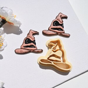 Op de afbeelding: Drie koekjesvormen in de vorm van de Sorteerhoed uit de Harry Potter-serie. De vormen zijn gemaakt van plastic en zijn in verschillende tinten bruin en zwart.