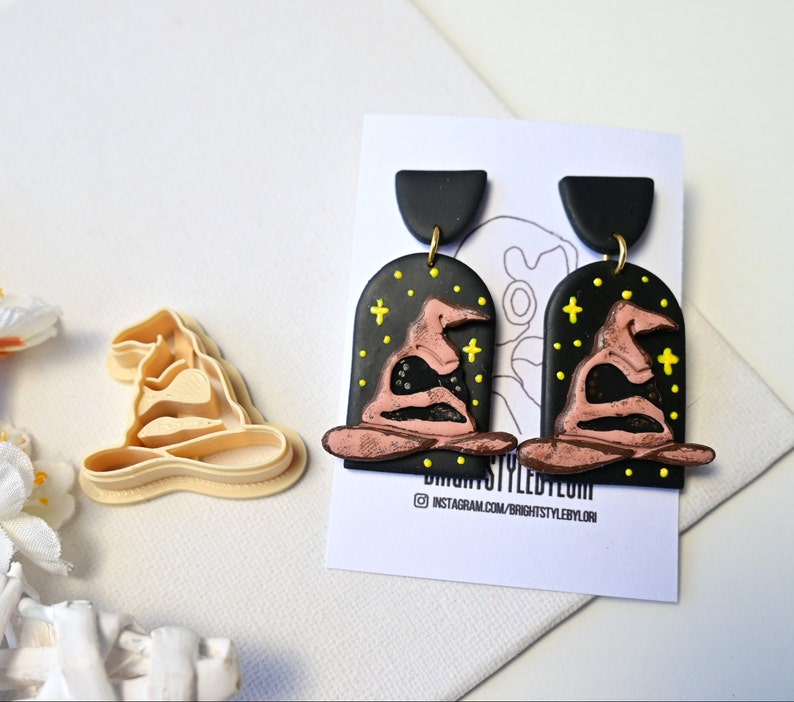 Op de afbeelding: Een paar zwarte en roze polymer clay oorbellen in de vorm van de Sorteerhoed uit de Harry Potter-serie. De oorbellen hebben een zwarte achtergrond met gele sterren en een roze Sorteerhoed met een zwarte rand. De oorbellen hangen aan zwarte haken.
