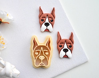 May include: Three cookie cutters shaped like boxer dog faces. Two are brown and white, and one is a beige plastic cutter.