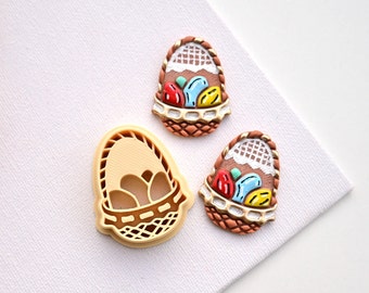 May include: Two ceramic Easter egg ornaments with a basket design and a plastic mold for making the ornaments. The ornaments are painted with brown, white, red, yellow, and blue details. The mold is beige.