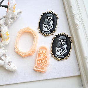 Op de afbeelding: Een verzameling handgemaakte gothic-thema sieraden en knutselbenodigdheden. De afbeelding toont twee zwarte en gouden ovale hangers met een meisje en kat ontwerp, samen met een frame en een uitsteker in perzikkleur.
