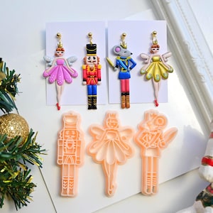 May include: Four dangle earrings featuring polymer clay figures of a fairy in a pink dress, a nutcracker, a mouse soldier, and a fairy in a yellow dress, displayed alongside three peach-colored plastic cookie cutters shaped like the same characters.