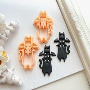 Op de afbeelding: Vier katvormige koekjessnijders. Twee zijn perzikkleurig en twee zijn zwart. Elke kat heeft een skeletontwerp en vleermuisvleugels. De snijders liggen op een wit oppervlak, met een witte decoratieve lijst op de achtergrond.