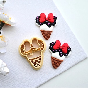 Può includere: Due stampini per biscotti a forma di cono gelato, uno è un cono semplice e l'altro è decorato con un fiocco rosso, glassa bianca e granella di zucchero viola.