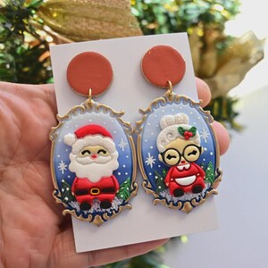K&ouml;nnte beinhalten: Ein Paar festliche Weihnachts-Ohrringe mit Weihnachtsmann und Frau Claus. Jeder Ohrring hat einen roten Aufsatz und einen goldfarbenen Rahmen. Der Weihnachtsmann tr&auml;gt einen roten Anzug und Hut, Frau Claus ein rotes Kleid und eine Brille.