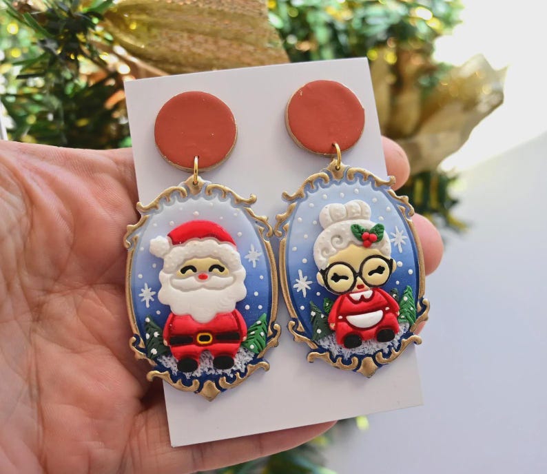 K&ouml;nnte beinhalten: Ein Paar festliche Weihnachts-Ohrringe mit Weihnachtsmann und Frau Claus. Jeder Ohrring hat einen roten Aufsatz und einen goldfarbenen Rahmen. Der Weihnachtsmann tr&auml;gt einen roten Anzug und Hut, Frau Claus ein rotes Kleid und eine Brille.