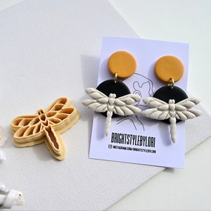 Op de afbeelding: Een paar oorhangers met een wit libellenontwerp met zwarte en mosterdgele accenten. De oorbellen zijn handgemaakt en hebben een unieke, speelse stijl.