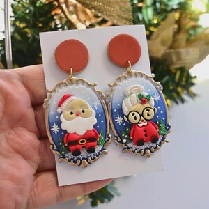 K&ouml;nnte beinhalten: Ein Paar Ohrringe mit Weihnachtsmotiv. Ein Ohrring zeigt den Weihnachtsmann, der andere Frau Claus. Beide Ohrringe haben einen blauen Hintergrund mit goldenem Rahmen und einem rostfarbenen Kreis.
