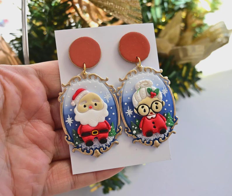 K&ouml;nnte beinhalten: Ein Paar Ohrringe mit Weihnachtsmotiv. Ein Ohrring zeigt den Weihnachtsmann, der andere Frau Claus. Beide Ohrringe haben einen blauen Hintergrund mit goldenem Rahmen und einem rostfarbenen Kreis.
