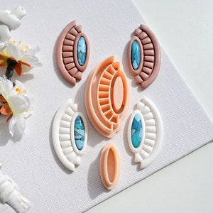 May include: A set of six clay earrings in various shapes and colours. The earrings are in the shape of half circles and have a turquoise stone in the centre. The earrings are on a white surface.