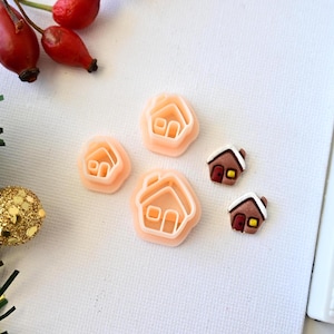 Puede incluir: Cuatro cortadores de galletas de plástico rosa en forma de casas, y dos galletas de fondant marrón en forma de casas con detalles rojos y amarillos.
