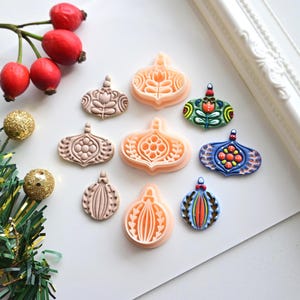 Pode incluir: Uma coleção de enfeites de Natal decorativos em várias formas e cores. Alguns são rosa claro, enquanto outros são pintados com intrincados desenhos florais em verde, azul e laranja. Os enfeites são dispostos sobre uma superfície branca com decorações festivas.