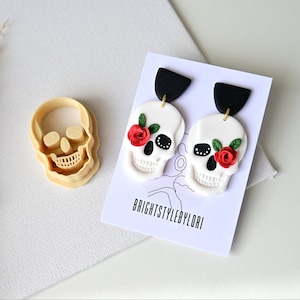 May include: A pair of white skull earrings with red roses and green leaves. The earrings are on a white card with the text "brightstylebylori".