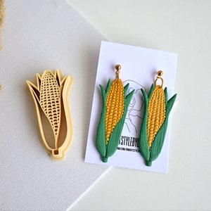 Könnte beinhalten: Ein Paar goldene Mais-Ohrringe mit grünen Blättern. Die Ohrringe sind aus Polymer Clay gefertigt und ein lustiges und verspieltes Accessoire.