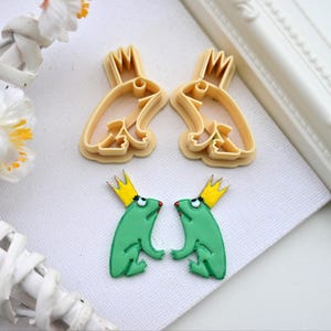 May include: Two green frog cookie cutters with gold crowns and red eyes. Two matching frog cookie cutters are shown in the background.
