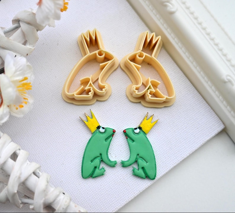 May include: Two green frog cookie cutters with gold crowns and red eyes. Two matching frog cookie cutters are shown in the background.