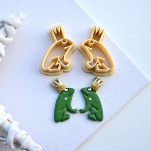 May include: Two green frog figurines with gold crowns, and two cookie cutters in the shape of frogs with crowns. The frogs are facing each other.