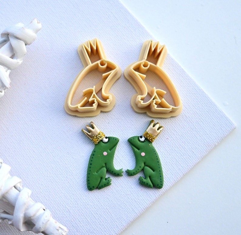 May include: Two green frog figurines with gold crowns, and two cookie cutters in the shape of frogs with crowns. The frogs are facing each other.