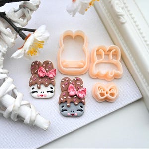 Könnte beinhalten: Zwei braune und graue fondant-förmige Hasendekorationen mit rosa Schleifen und Schokoladenüberzug. Es gibt drei Plastik-Ausstecher in Form eines Hasen, einer Schleife und eines Hasen mit einer Schleife.