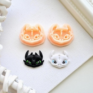 Könnte beinhalten: Vier Drachen-Bastelstücke. Zwei sind hellorangefarbene Formen, und die anderen beiden sind fertige Drachengesichter. Eines ist schwarz mit grünen Augen, und das andere ist weiß mit blauen Augen. Die Teile liegen auf einer weißen, strukturierten Oberfläche.