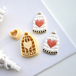 Op de afbeelding: Twee witte keramische vogelkooi-versieringen met gouden rand en een roze hartontwerp. De ornamenten liggen naast twee beige plastic koekjesvormen in de vorm van een vogelkooi en een hart.