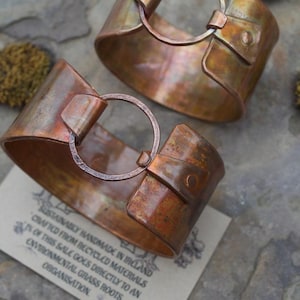 May include: Two wide copper cuff bracelets with a weathered, oxidized finish. Each bracelet features a central circular ring connector attached to the band with a folded metal tab and a single rivet. The metal has varied brown and orange tones.