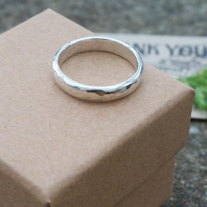 May include: A silver ring with a textured, hammered finish. The band has a rounded profile and a reflective metallic surface. The ring is positioned on top of a square, light brown cardboard box, showcasing its simple and clean design.