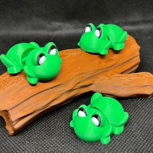 May include: A collection of green frog figurines with white and black eyes are arranged on a brown, textured log. One frog is positioned separately. The frogs are a bright green color, and the log has a natural wood grain appearance.