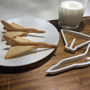May include: Two light brown cookies shaped like fighter jets on a white plate.  A white plastic cookie cutter shaped like a fighter jet is on a wooden surface.
