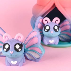 Puede incluir: Una figura de mariposa impresa en 3D en tonos morado, rosa y azul. La mariposa tiene ojos grandes y expresivos y un detalle en forma de corazón. Una estructura decorativa a juego está en el fondo, todo sobre un fondo rosa suave.